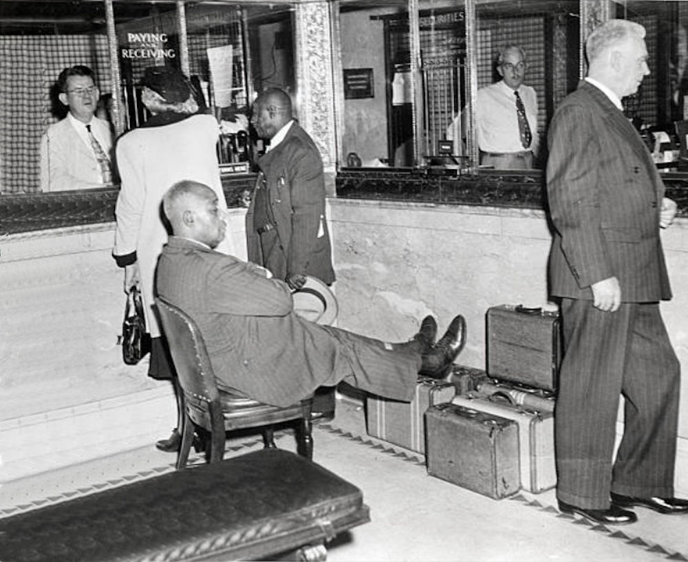 Standing at the window holding the straw hat
10/21/1949- Newark, NJ - One of Father Divine's followerskeeps his eyes, and rests his feet, on nearly $500,000 which he and other members carried to the bank in payment for the Hotel Riviera.

